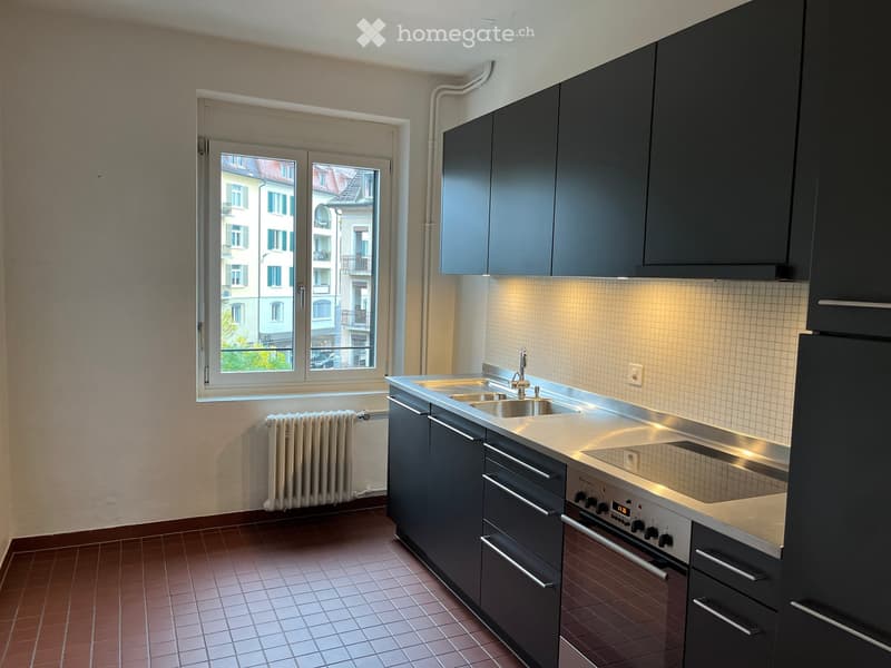 Modern kitchen with dark grey cabinets, stainless steel sink and oven, tiled floor, and a window overlooking a cityscape.