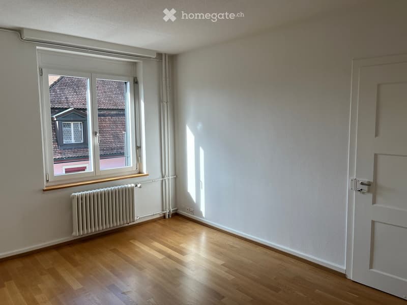 Empty room with wooden flooring, white walls, a window with a radiator below, and a door on the right side.