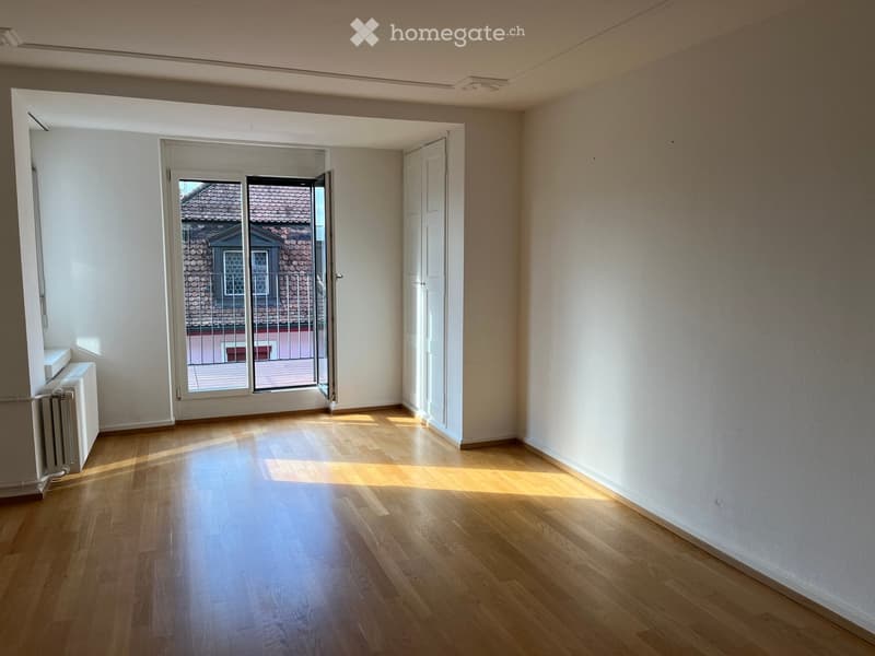 Empty room with wooden flooring, white walls, and a large window with a view of a neighboring building. The room is well-lit with natural light.