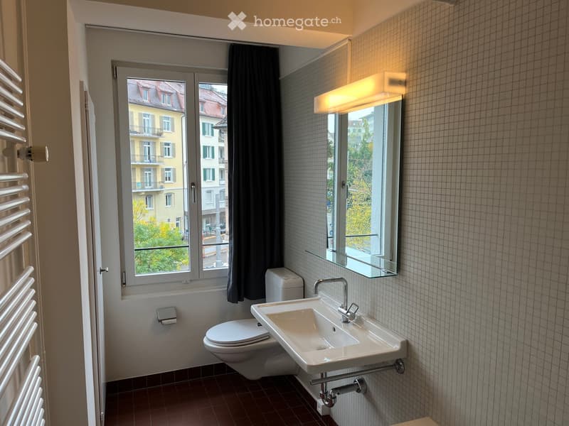 Modern bathroom with a toilet, sink, and mirror. The room features tiled walls and a window with a view of the city.
