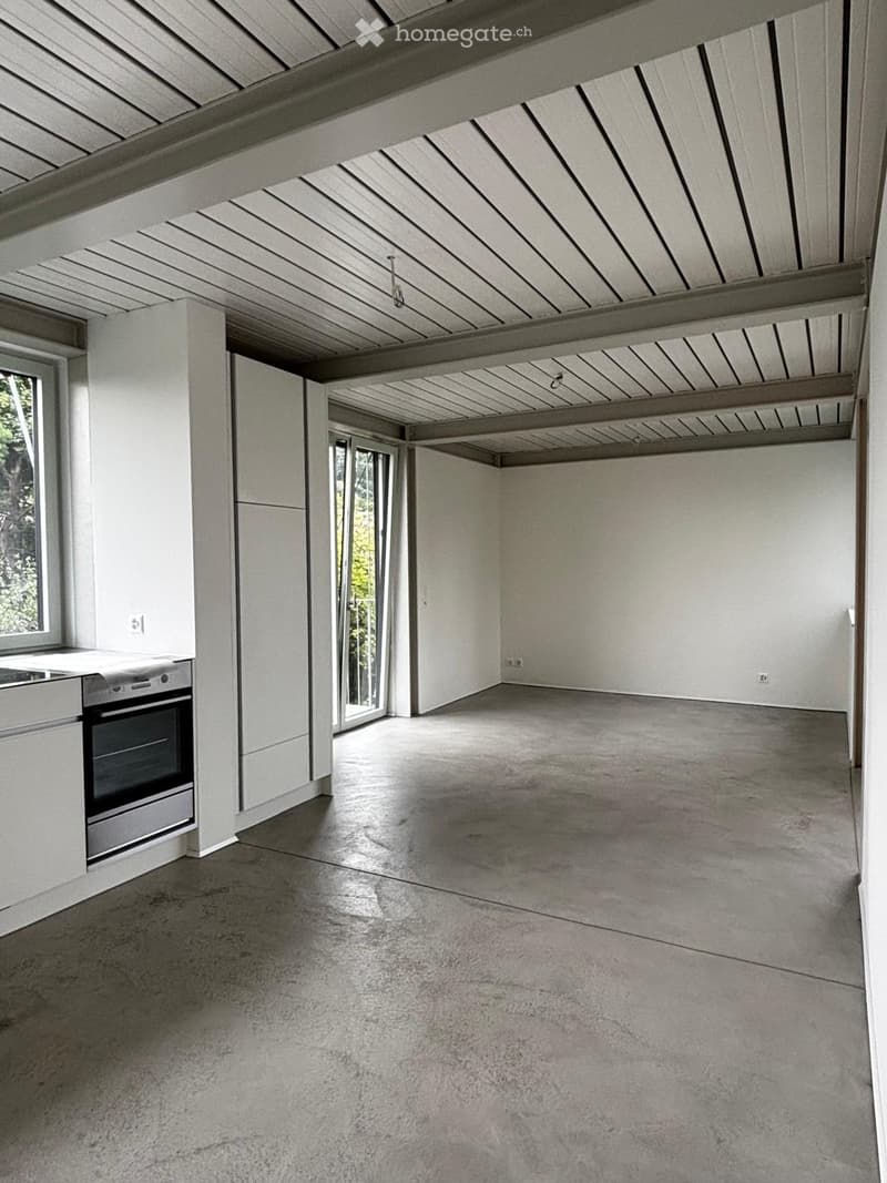 An unfurnished room with a concrete floor, white walls, and a modern kitchen area featuring a built-in oven and countertop. The room has large windows and a high, exposed wooden ceiling structure.