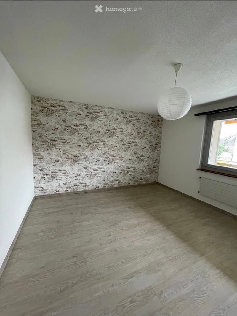 Empty room with wooden flooring, exposed brick wall, white pendant light, and a window with a view.