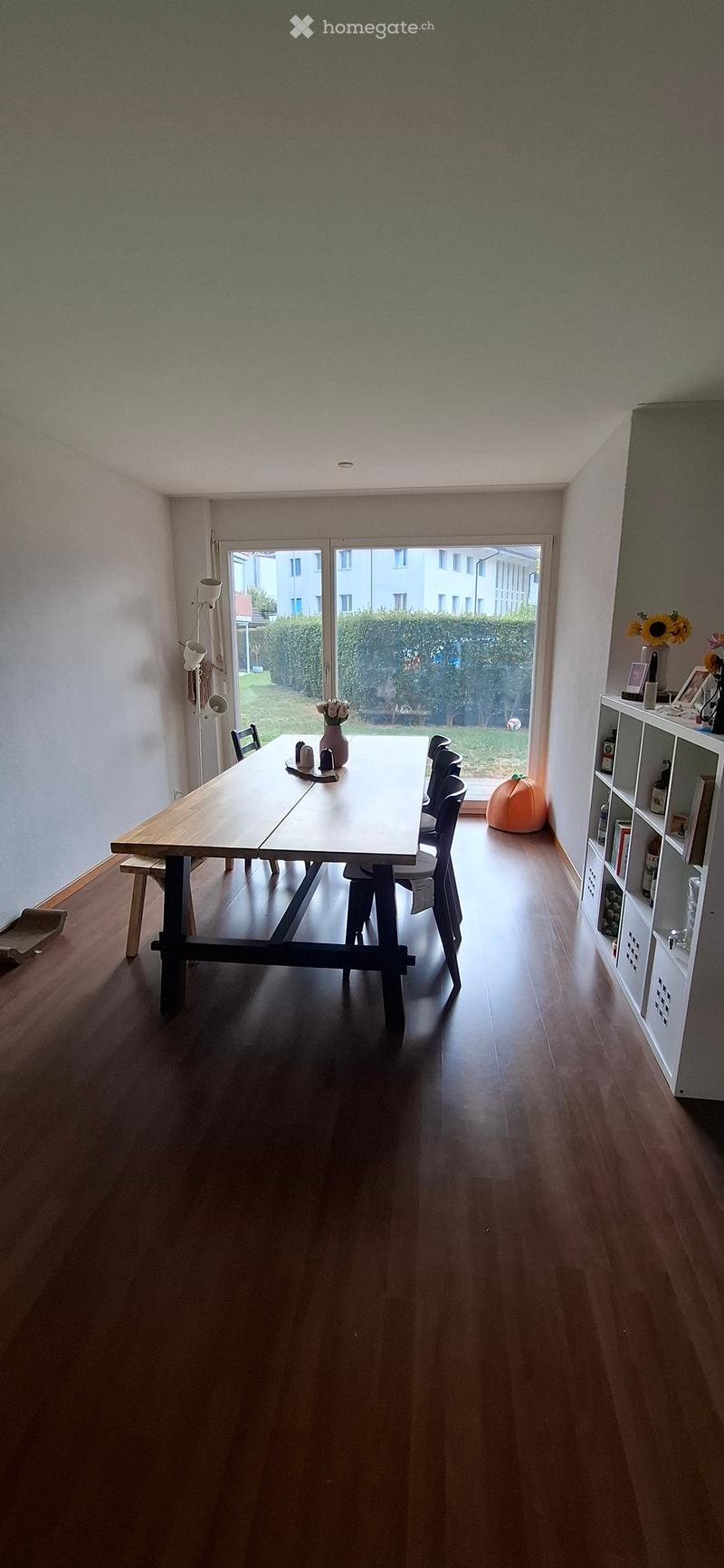 Dining area with wooden table, chairs, vase, shelf, and pumpkin near the window.
