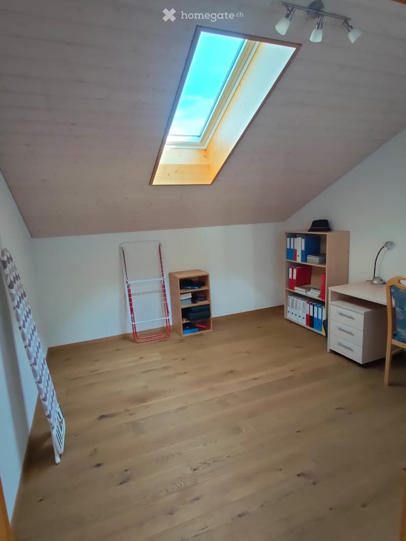Bright attic room with wooden flooring, slanted ceiling, skylight window, and modern furniture including a desk, chair, bookshelf, and small cabinet.
