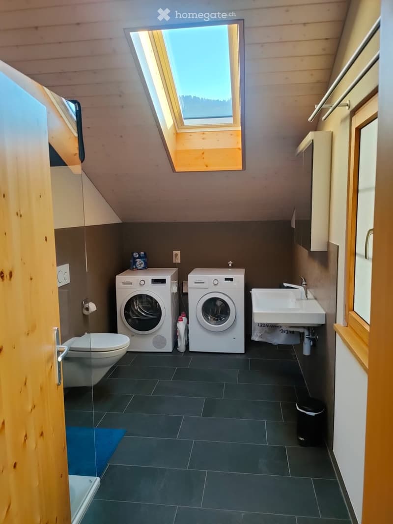 Modern bathroom with dark tiled floor, white washing machine and dryer, toilet, sink, and wooden ceiling with skylight windows.