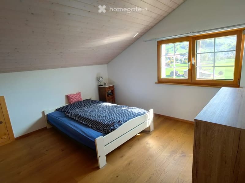 Cozy attic bedroom with wooden floors, slanted wooden ceiling, large windows offering a scenic view, a single bed with blue and black bedding, a nightstand with a lamp, and a small wooden table.