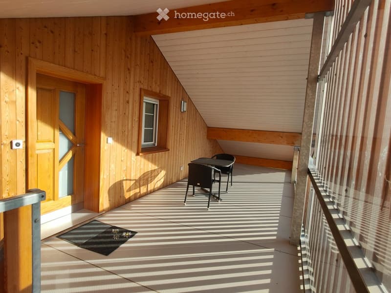 Wooden balcony with a small table and two chairs, wooden walls, a window, and a doormat.