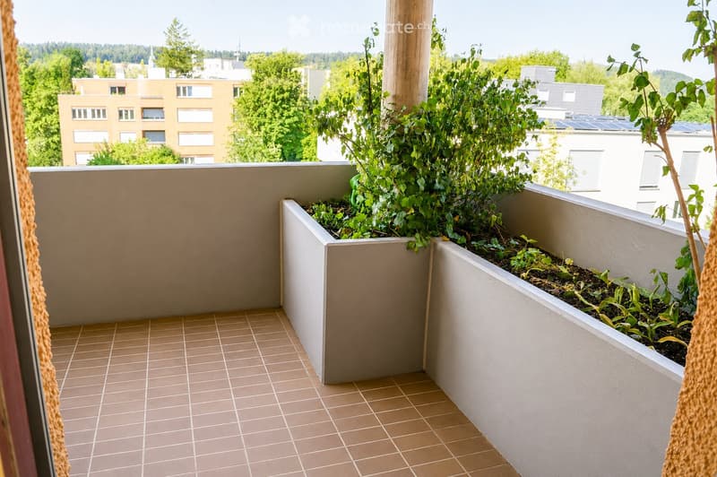 Balcony with tiled floor, raised planters containing green plants, and a view of surrounding buildings and greenery.