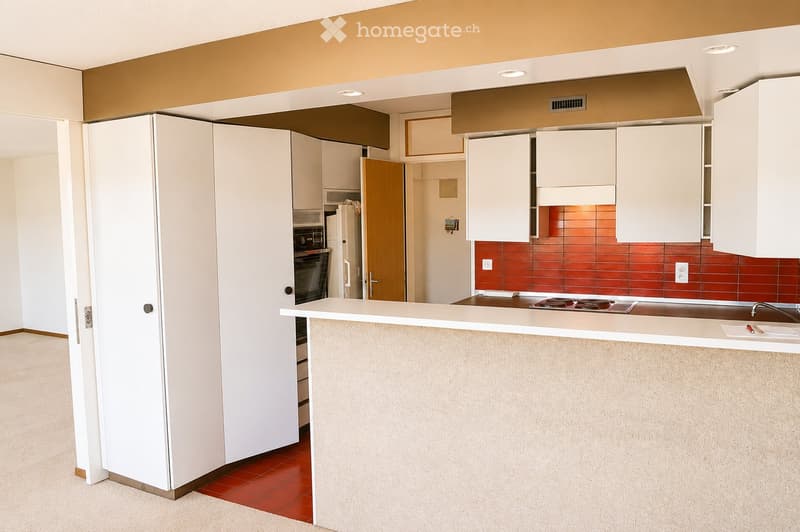Modern kitchen with white cabinets, a central island counter, and a stove. The floor is tiled in red, and the walls feature a mix of white and red tiles.