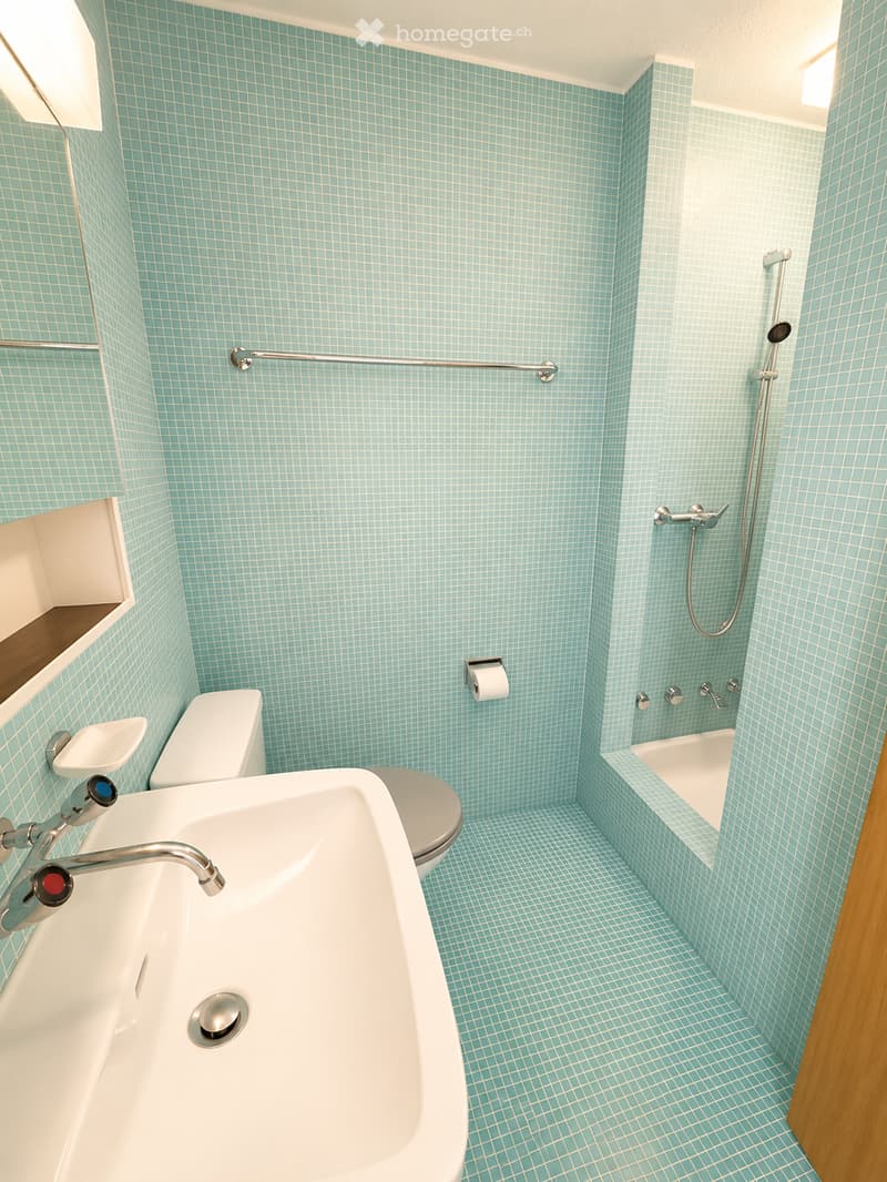 Modern bathroom with light blue tiled walls and floor, featuring a white sink, toilet, bathtub, and a handheld showerhead with a rail.