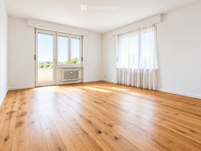 Empty room with light wooden flooring, white walls, large windows with sheer curtains, and a sliding glass door leading to a balcony.