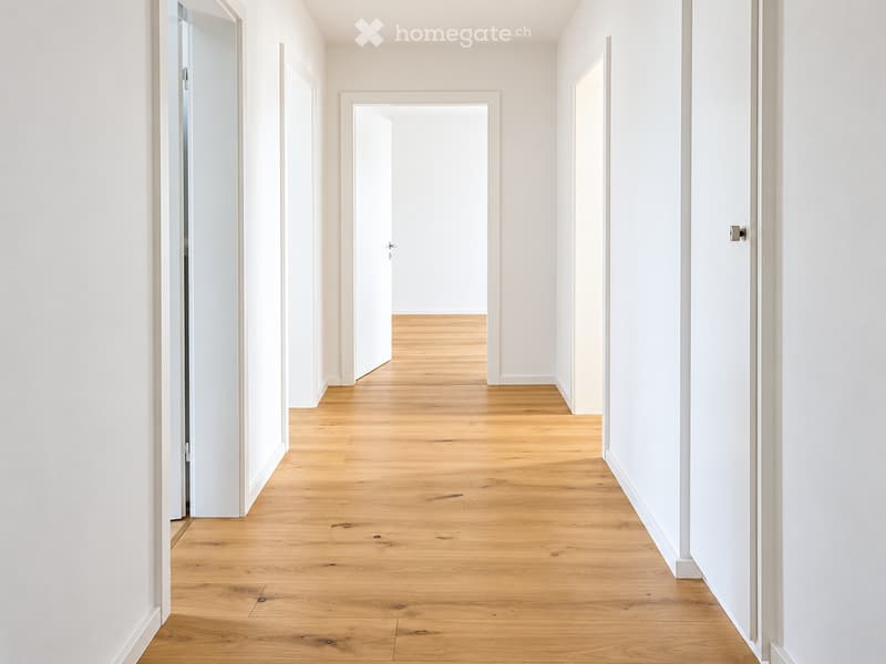 Empty hallway with light wooden flooring and white walls, featuring several doors and a glass-paneled door on the left.
