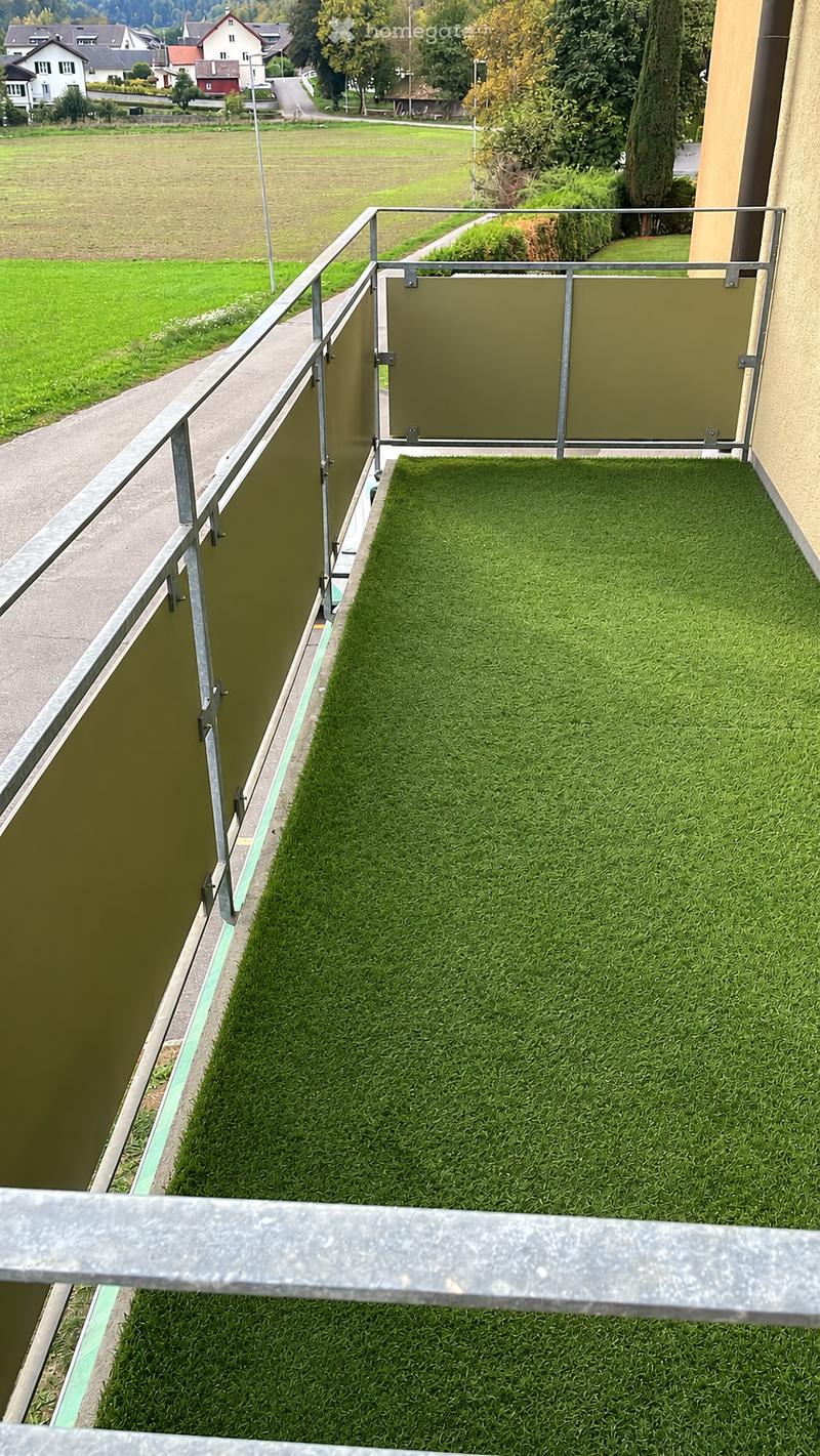 Balcony with artificial grass surface, metal railing, and a view of a rural landscape with fields and houses.
