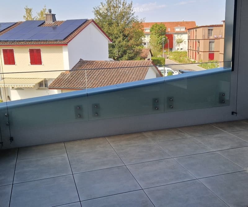 Balcony with tiled floor, glass railing, and a view of neighboring houses and trees.