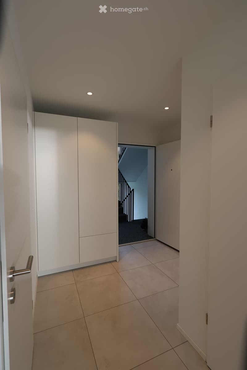 A clean, modern hallway with tiled flooring, white walls, and recessed ceiling lights. A white cabinet is on the left, and an open doorway leads to a staircase area.
