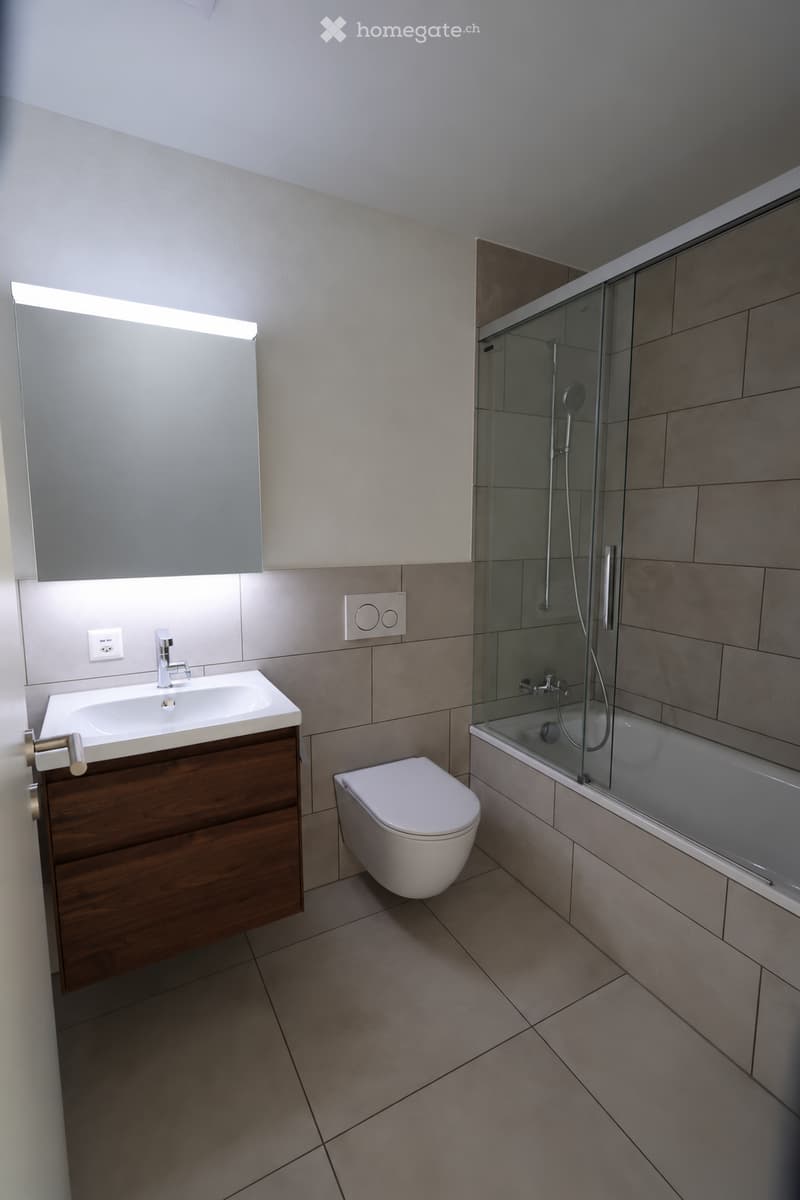 Modern bathroom with a wooden vanity, sink, toilet, bathtub with glass enclosure, and a mirror with built-in lighting.