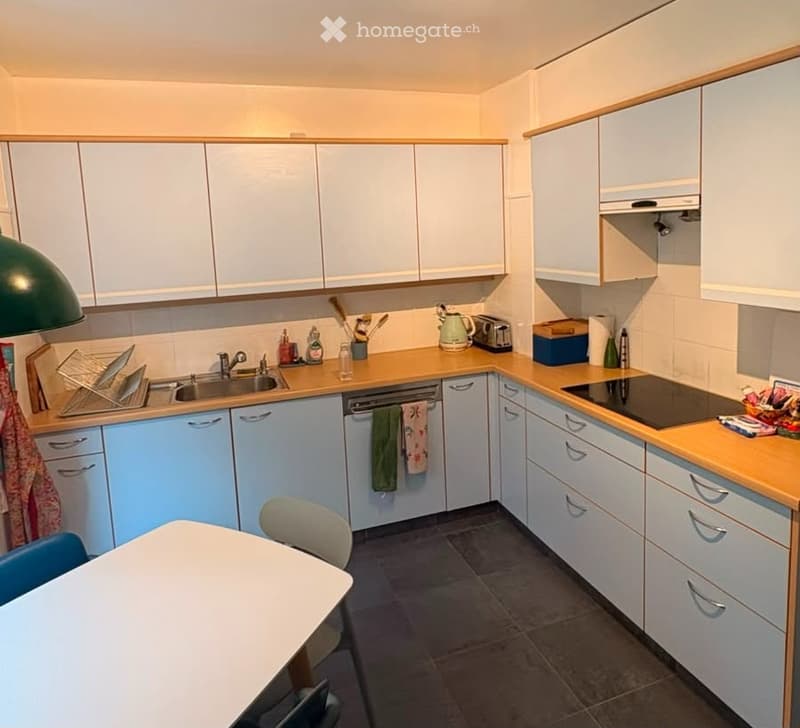 Bright kitchen with light blue cabinets, wooden countertops, and stainless steel appliances including a dishwasher and induction stove. Features a sink, faucet, and a small dining area with a table and chairs.