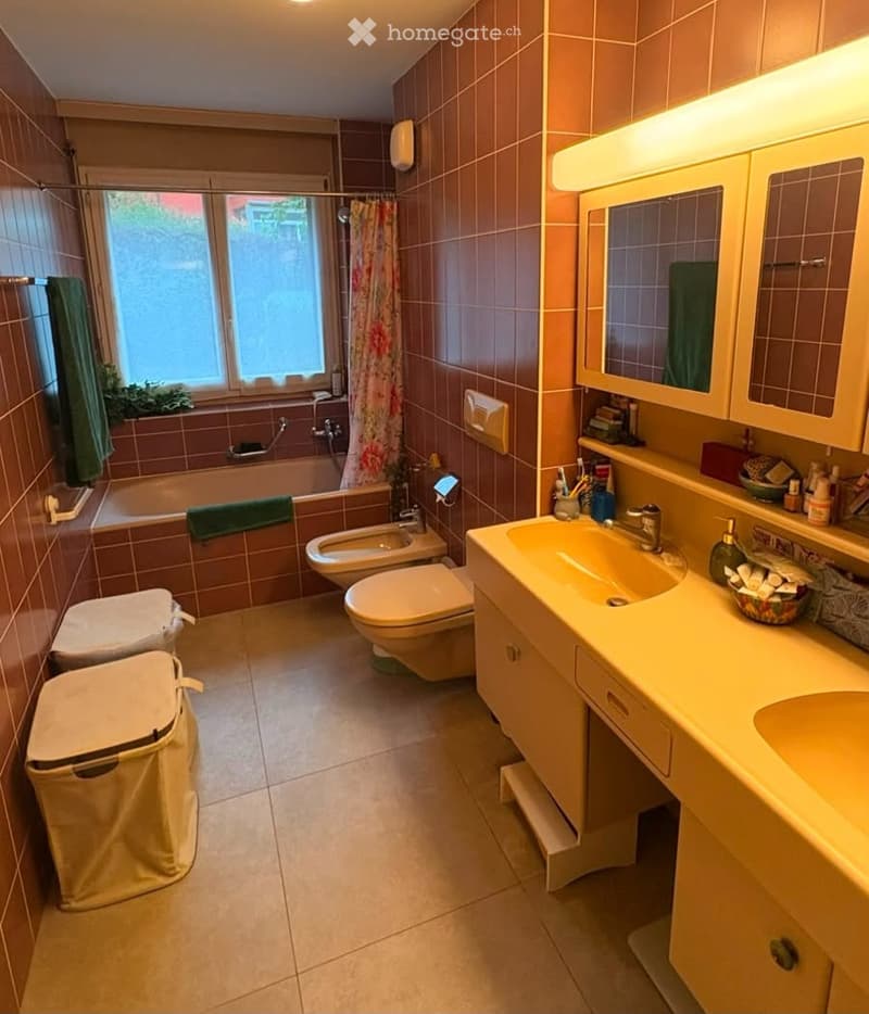 Bathroom with brown tiled walls and floor, featuring a bathtub, toilet, bidet, sink with mirror and cabinet, and a window with a shower curtain.