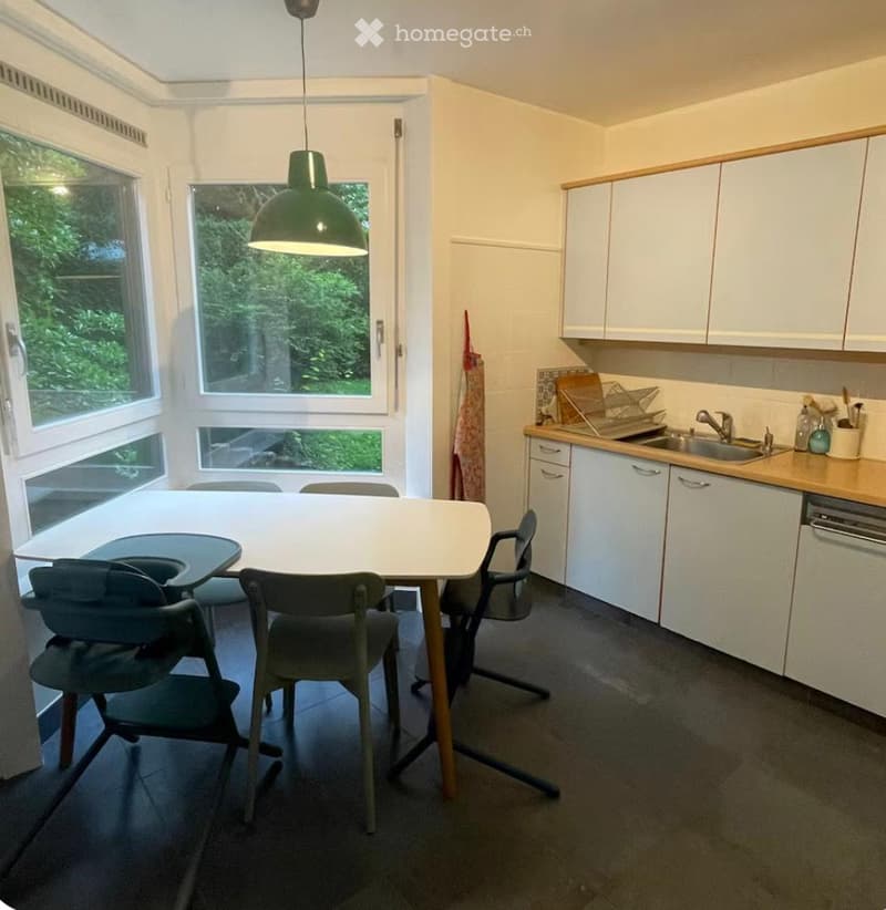 Bright kitchen and dining area with white cabinets, a central dining table with chairs, and a sink area with a faucet and dishwasher. Large windows provide natural light.