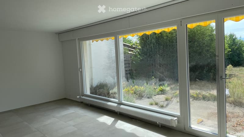 Empty room with large windows and a radiator, showing a view of a garden area outside.