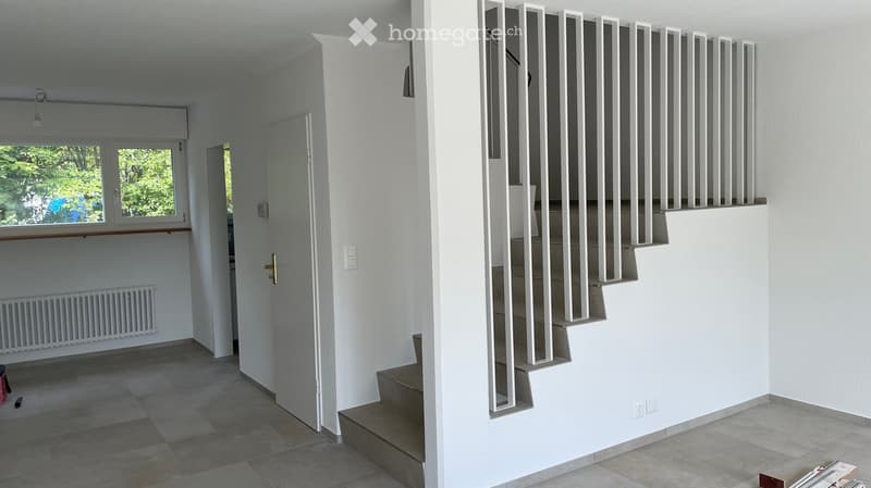 Bright, modern hallway with tiled flooring, white walls, and a staircase leading upwards. Features include a window with a view of greenery, a radiator, and a door leading to another room.