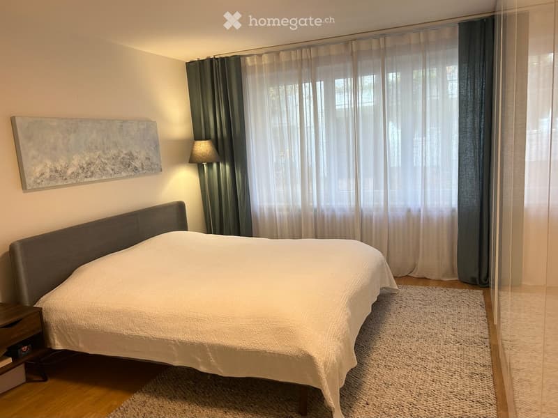 Modern bedroom with a large bed, nightstand, lamp, and large window with curtains. A painting is on the wall, and a small rug is on the floor.