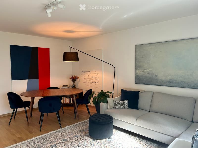 Modern living room with a grey sofa, ottoman, wooden dining table, chairs, floor lamp, and decorative artwork.