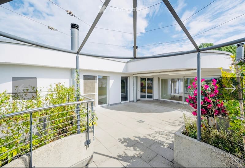 Modern outdoor patio area with white walls, large glass doors, and lush greenery, including flowering plants and shrubs.
