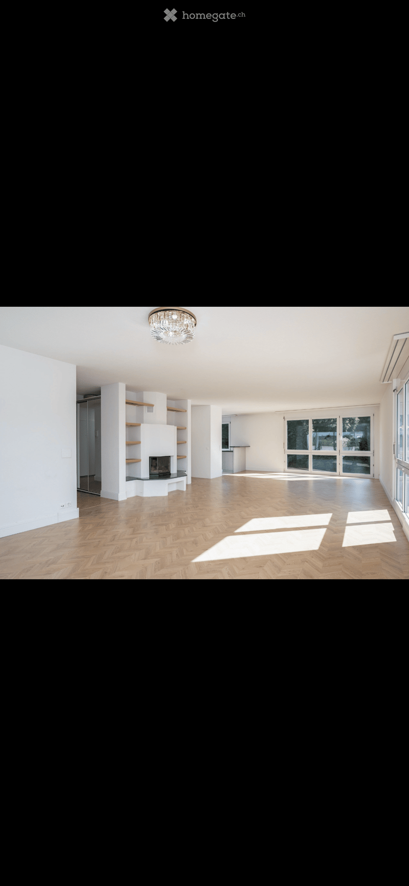 Empty room with wooden herringbone flooring, white walls, built-in fireplace with shelves, large windows, and a chandelier.