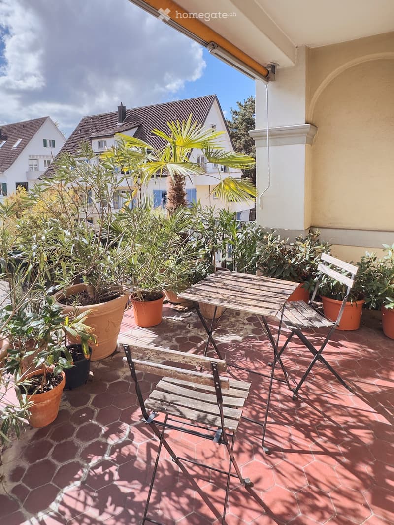 Balcony with wooden table and chairs, surrounded by potted plants and greenery, overlooking a residential area.