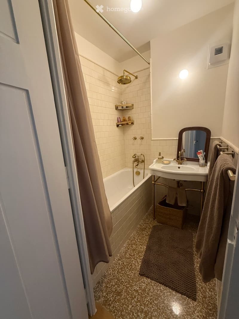 Cozy bathroom with a bathtub, sink, mirror, and showerhead. Features a decorative shelf, towel, and bathmat on a patterned floor.