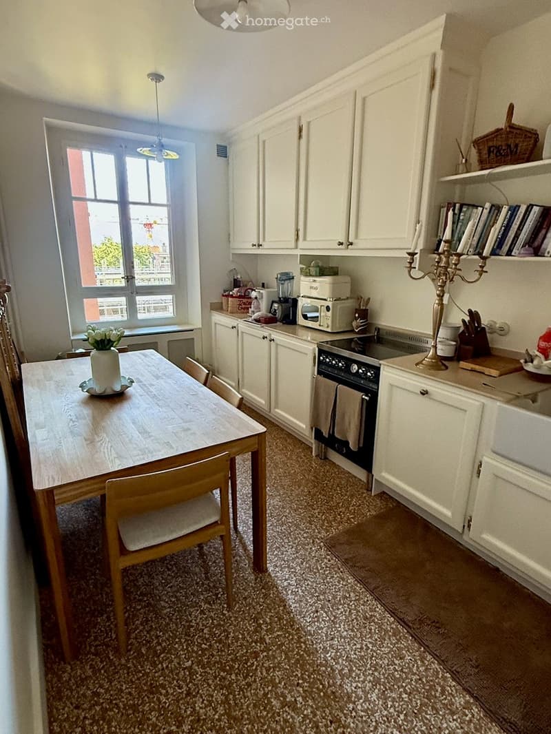 Bright kitchen with white cabinets, a wooden dining table with chairs, and various appliances including an oven, microwave, and blender. A window offers natural light, and decorative items such as a candle holder and a flower vase are present.