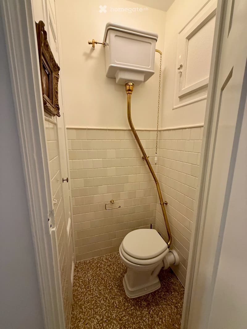 A small bathroom with white tiled walls, a speckled floor, a toilet, and a toilet paper holder with a gold chain.