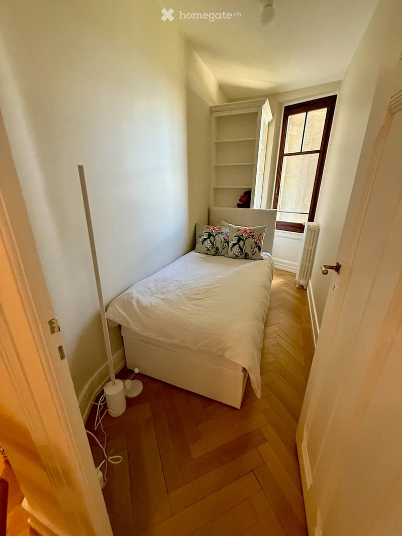 Cozy bedroom with a single bed, pillows, a small bookshelf, radiator, and a floor lamp. Wooden flooring and white walls.