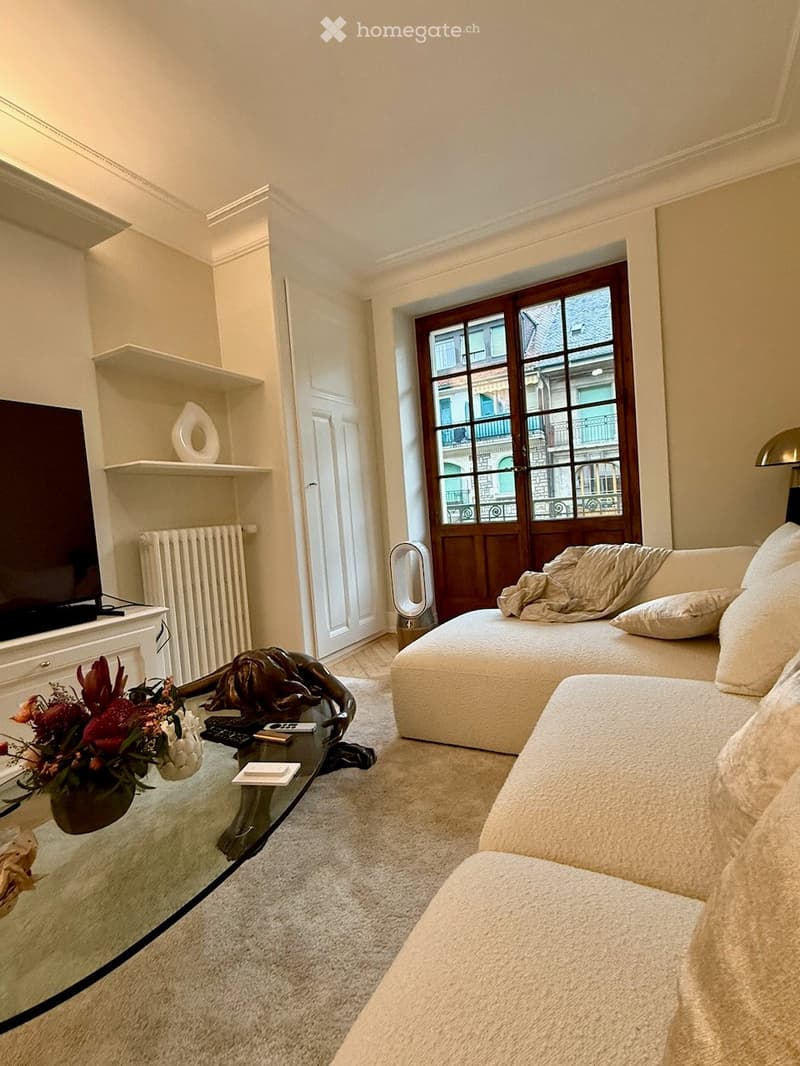 Modern living room with a white sofa, glass coffee table, decorative sculpture, flower arrangement, TV on a white cabinet, and large windows with wooden frames.