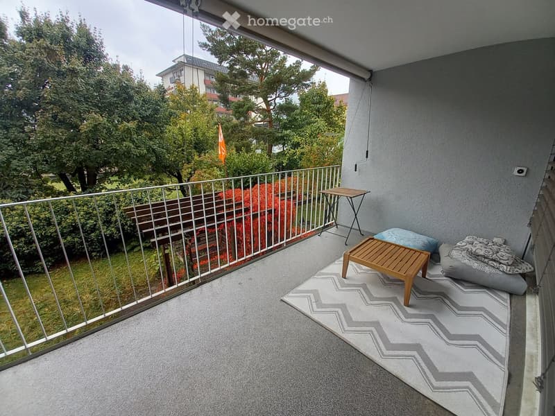Balcony with a rug, small wooden table, stool, and two poufs, overlooking a green garden area.