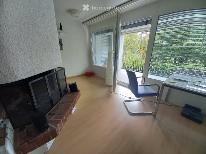Bright room with wooden flooring, fireplace, large windows, balcony access, chair, table, and decorative vase.