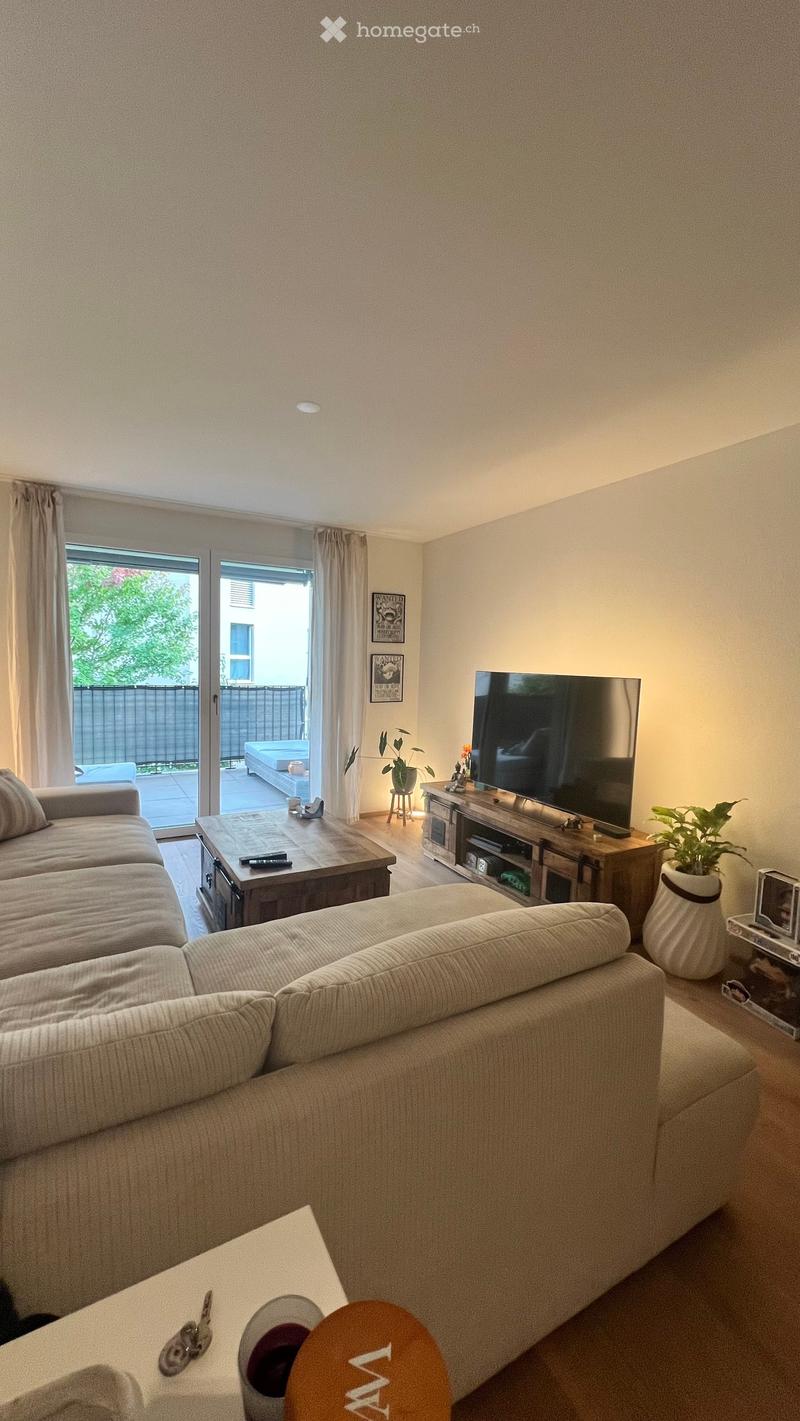 Modern living room with a large sectional sofa, coffee table, flat-screen TV on a wooden stand, and a small plant in a pot. The room features large windows with curtains and a sliding glass door leading to a patio.