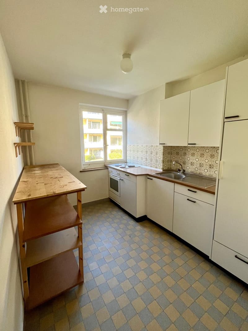 Compact kitchen with white cabinets, a sink, an oven, and a wooden table. The room features a checkered tile floor and a window offering a view of the building outside.