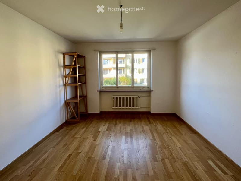 Empty room with wooden flooring, white walls, a window with a radiator below, and a simple wooden shelving unit in the corner.