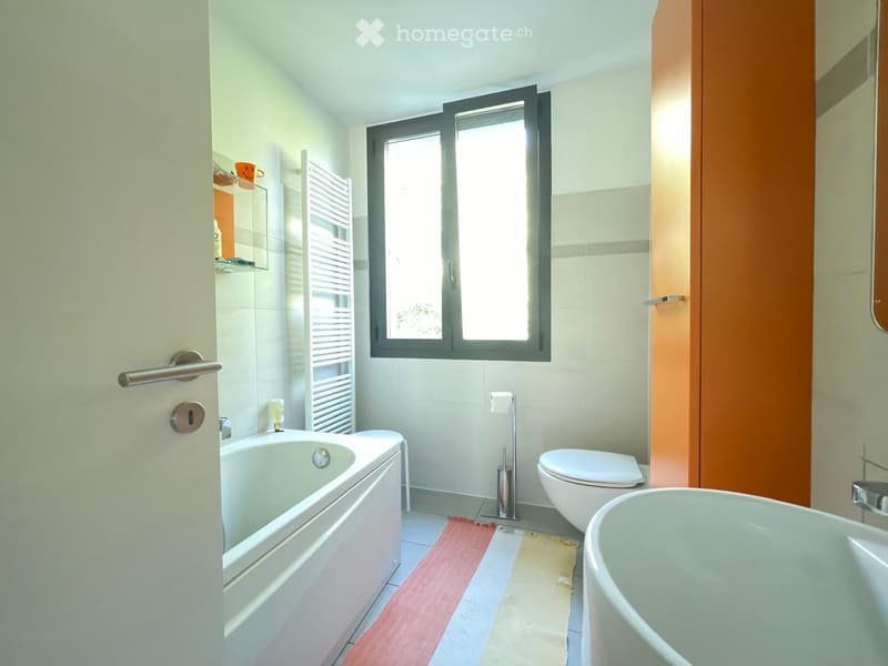 Modern bathroom with white tiles, a bathtub, toilet, towel rack, and a window letting in natural light.