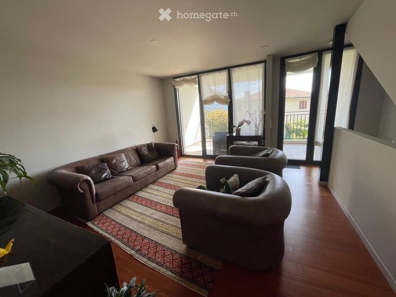 Modern living room with wooden flooring, large windows, and a balcony. Features a brown leather sofa, armchair, coffee table, and decorative plants.
