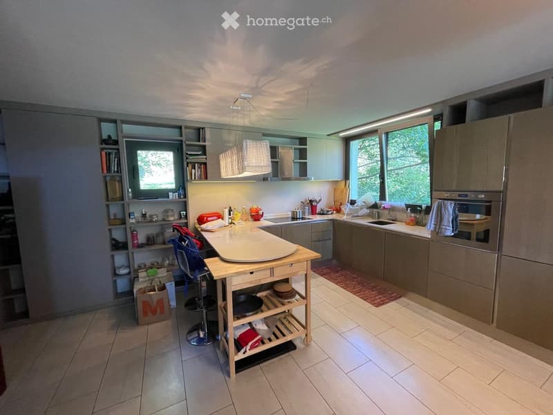 Modern kitchen with light-colored cabinets, a central island, and various appliances including a sink, oven, and refrigerator. Shelves hold books and kitchen items, and a small table with a chair is present.