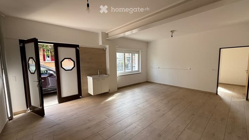 Empty room with wooden flooring, white walls, and a small sink area. The room has large windows and a door leading outside.