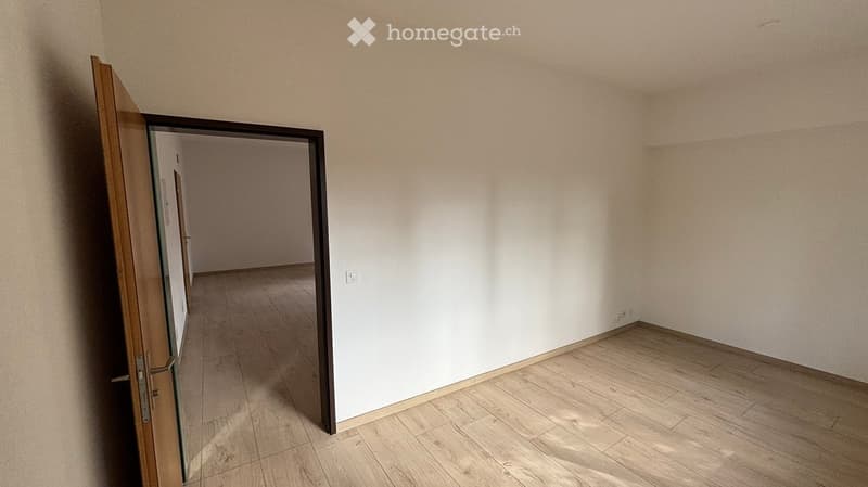 Empty room with light wooden flooring and white walls, featuring an open doorway leading to another room.