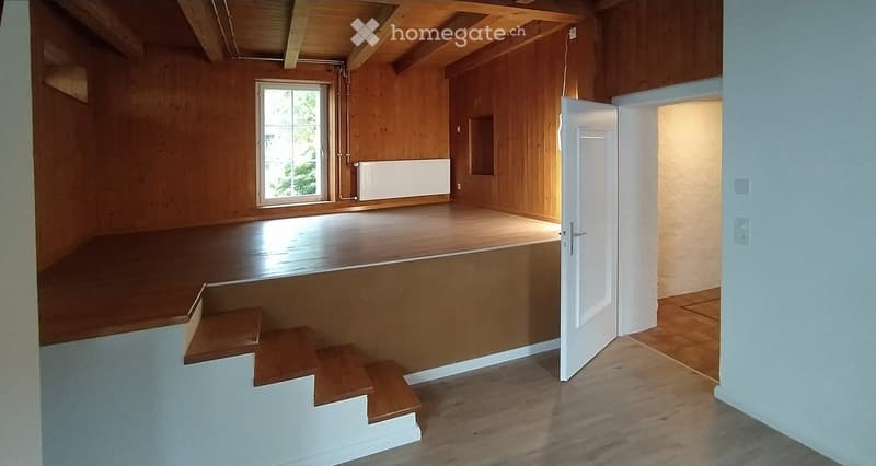 Empty room with wooden walls, wooden floor, a window, a radiator, and a door leading to another room.