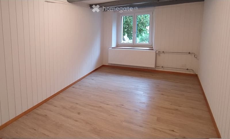Empty room with wooden flooring, white paneled walls, and a window with a radiator below it.