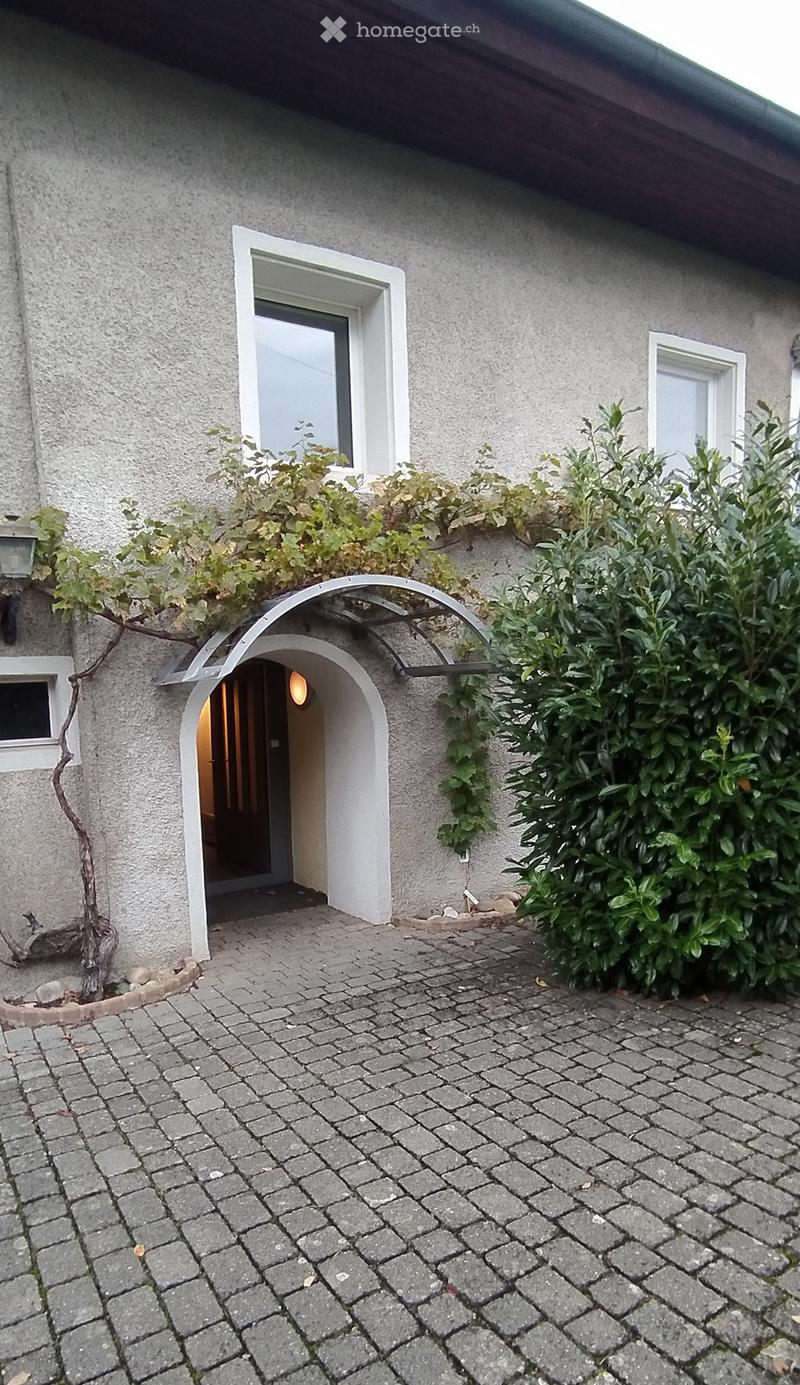 Exterior view of a two-story house with a gray facade, arched entrance, and surrounding greenery.