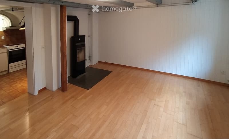 Empty room with wooden flooring, white walls, and a fireplace in the corner. Adjacent kitchen area visible with stove and cabinets.