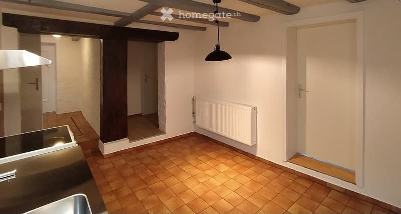 Empty room with wooden tiled floor, white walls, exposed wooden beams on the ceiling, radiator, and a hanging light fixture.